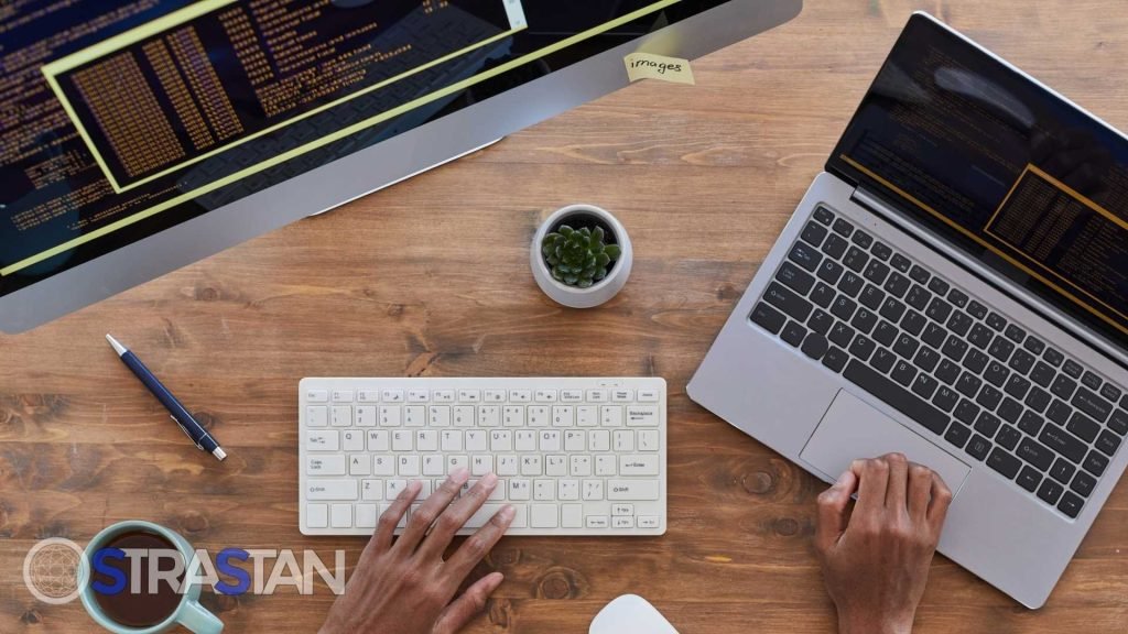An overhead view of a desk with a person typing on a keyboard, a laptop, a desktop monitor displaying code, a small plant, a pen, and a cup of coffee.