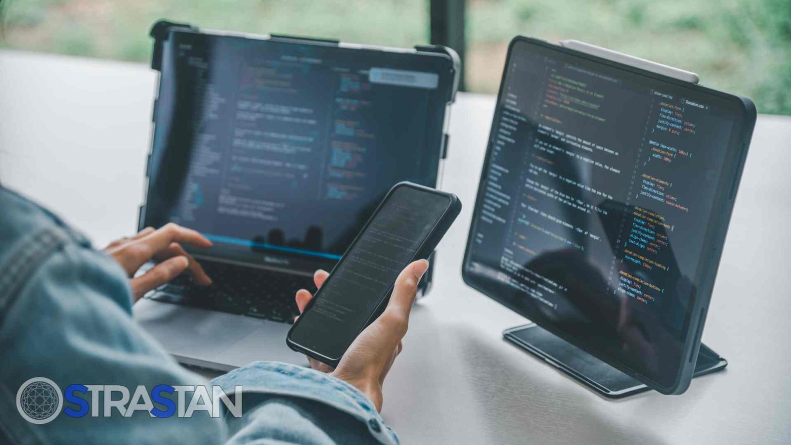A person holds a smartphone with a laptop and a tablet in the background, all displaying lines of code, symbolizing the development process for both mobile vs web apps.